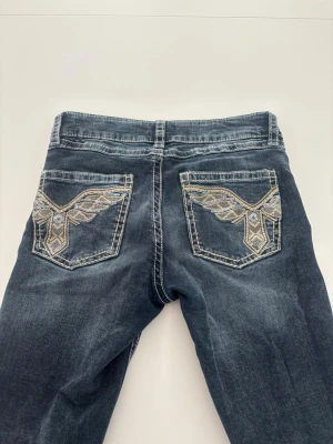 Mörkblå jeans med broderade fickor från Gina Young  - Snygga mörkblå jeans med unika broderade detaljer i beige och silver på bakfickorna. Jeansen är low waist och bootcut. Mörkblå, mycket bra skick använda ett fåtal gånger