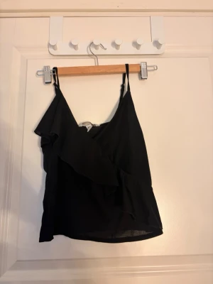 Svart linnetopp med volang - Snygg svart linnetopp med tunna axelband och volangdetalj framtill. Toppen har en luftig och lätt känsla, perfekt för varma dagar. Passar till både jeans och kjol för en trendig look.
