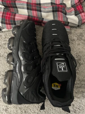Nike Air Vapormax Plus svarta sneakers - Svarta Nike Air Vapormax Plus sneakers med ikonisk bubbelsula i genomskinlig plast. Ovandelen är i mesh och syntet med vågiga detaljer och reflekterande inslag. Klassisk snörning och bekväm passform. Perfekta för dig som gillar streetwear och sportig stil.