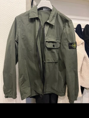 Grön overshirt från Stone Island - Snygg militärgrön overshirt från Stone Island med klassisk patch på ärmen. Jackan har dragkedja framtill, bröstficka med knapp och krage. Tillverkad i ett slitstarkt bomullsmaterial som ger en clean och stilren look.