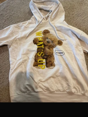 Vit hoodie med nalle och text - Säljer en vit hoodie med huva och dragsnören. På framsidan finns ett stort tryck av en söt brun nallebjörn, gul text som säger 'WHAT'S UP' och en pratbubbla med 'HI THERE!'. Hoodien har ribbade muddar och är perfekt för en avslappnad stil.