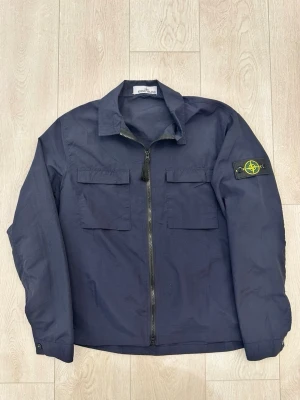 Mörkblå overshirt från Stone Island - Snygg mörkblå overshirt från Stone Island med dragkedja framtill, två bröstfickor och klassisk patch med logga på vänster ärm. Overshirten har en rak passform och är tillverkad i ett lätt, slitstarkt material. Perfekt för lager på lager och streetwear-stil.