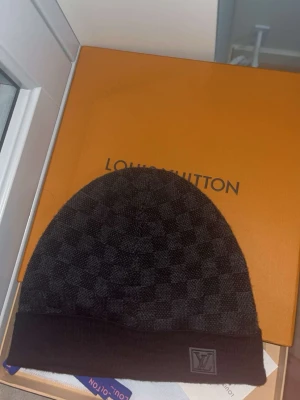Louis Vuitton svart rutig mössa - Säljer en snygg svart mössa från Louis Vuitton med diskret rutmönster och vikbar kant. Mössan har en liten LV-logga framtill och är tillverkad i mjukt material som känns lyxigt och stilrent. Perfekt accessoar för dig som gillar exklusiva detaljer.