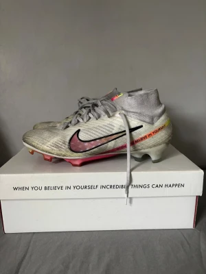 Nike Mercurial fotbollsskor vit/rosa (Limited edition!) - Säljer ett par Nike Mercurial fotbollsskor i vitt med rosa och svarta detaljer. Skorna har hög strumpkrageagig design och texten 'When you believe in yourself' på sidan. Sulan är delvis rosa och skon är tillverkad i syntetmaterial för bästa grepp och känsla på planen. Orginallåda följer med. Skicket är perfekt, inga trasiga delar, bara färgade ifrån konstgräset. Priset är ej fast, kan diskuteras!!