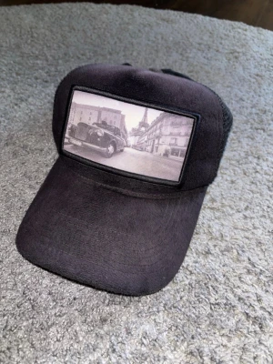 Svart keps HL2DRIP med fototryck - Säljer en svart keps från HL2DRIP med mesh på sidorna och ett stort fototryck framtill som visar en bil och Eiffeltornet i bakgrunden. Kepsen har böjd skärm och justerbar baksida för perfekt passform. Snygg och unik accessoar för dig som gillar streetwear.