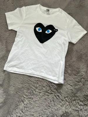 Vit Comme des Garçons Play t-shirt - Vit t-shirt från Comme des Garçons Play med ett stort svart hjärta och blå ögon tryckt på bröstet. Klassisk rund hals och korta ärmar. Perfekt för dig som gillar streetwear och vill sticka ut med en ikonisk look. 