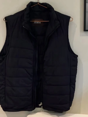 Svart dunväst från Oscar Jacobson - Stilren svart dunväst från Oscar Jacobson med hög krage och dragkedja framtill. Västen har quiltad design och två sidofickor. Perfekt för lager-på-lager och ger en clean look till din outfit. Köpt på NK i Göteborg och använts fåtal gånger. Skickas samma dag som köpt! 