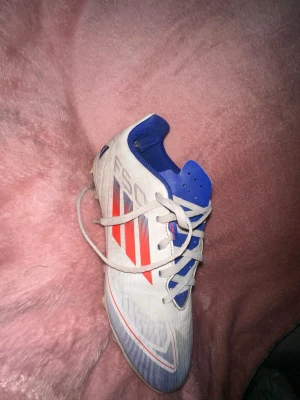 Adidas F50 vita och blå fotbollsskor - Säljer ett par Adidas F50 fotbollsskor i vitt med blå och röda detaljer. Skorna har klassiska tre ränder på sidan, snörning och dobbar för spel på gräs. Insidan är blå och ovandelen har en sportig, aerodynamisk design i syntetmaterial.