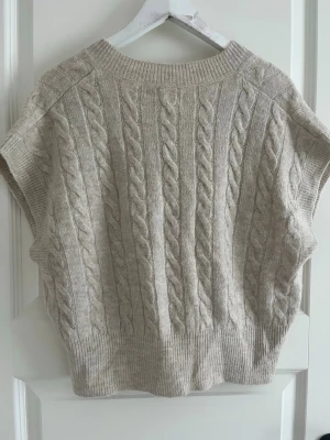 Beige stickad väst från ONLY - Säljer en beige stickad väst från ONLY i storlek XS. Västen har ett klassiskt flätstickat mönster, ribbade kanter och en rund halsringning. Perfekt att styla över en skjorta eller t-shirt för en trendig lager-på-lager-look.