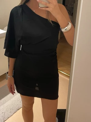 Svart oneshoulder kortklänning - Snygg svart kortklänning med oneshoulder-design och lös ärm på ena sidan. 