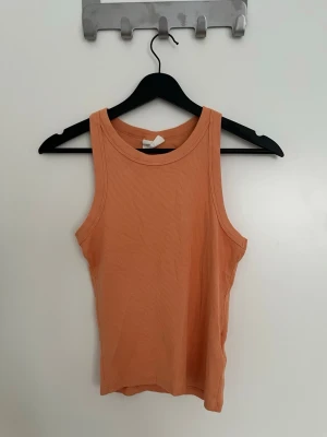 Ribbad persikoorange linnetopp från Gina Tricot - Säljer en ribbad, croppad linnetopp i en snygg persikoorange färg från Gina Tricot. Toppen är ärmlös med rund halsringning och sitter tight och mjukt mot kroppen. .