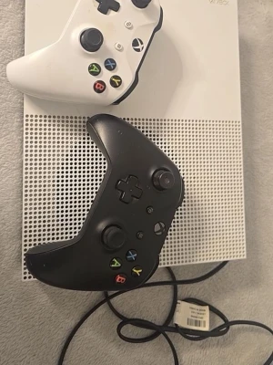 Xbox One S med två handkontroller - Säljer en Xbox One S i vit färg med två trådlösa handkontroller, en svart och en vit. Konsolen har en stilren design med ventilerat mönster på ovansidan och klassiska Xbox-knappar på kontrollerna. Perfekt för gaming med vänner!