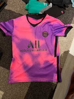 PSG Mbappé matchtröja rosa/lila - Säljer en PSG matchtröja i rosa och lila med svart V-ringning och detaljer. Framsidan har klubbmärke och sponsorlogga, baksidan har Mbappé och nummer 7. Tillverkad i lätt och ventilerande funktionsmaterial, perfekt för fotbollsträning eller match. Dock har trycket där bak gått av men man ser vad det står. Storlek S men passar även M