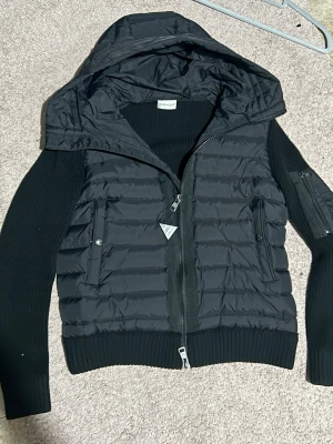 Svart pufferjacka med stickade ärmar - Snygg svart pufferjacka från Moncler med quiltad front och huva. Ärmarna är stickade i svart material och har en ficka med dragkedja på ena ärmen. Jackan har två framfickor med tryckknappar och dragkedja framtill. Perfekt för kalla dagar.