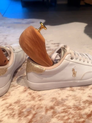 Vita sneakers från Polo Ralph Lauren - Snygga vita sneakers från Polo Ralph Lauren med guldfärgade detaljer och logga på sidan. Skorna har lågt skaft, vita snören och glittrig gulddetalj på hälen. Tillverkade i slätt konstläder för en clean look. Endast använda vid konfirmation.