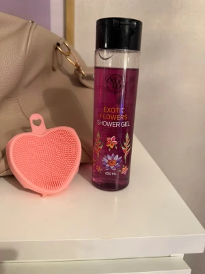Exotic Flowers duschgel & silikonborste - Säljer en Exotic Flowers Shower Gel från IDC Institute, 250 ml, i en genomskinlig flaska med lila gel och blommigt tryck. Medföljer gör en hjärtformad, rosa silikonborste perfekt för rengöring av huden. Perfekt för dig som vill ha en fräsch och blommig duschrutin.