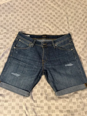 Blå jeansshorts med slitningar - Säljer ett par klassiska blå jeansshorts med uppvikta benslut och slitna detaljer framtill. Shortsen har fem fickor och gul kontrastsöm. Perfekta för en avslappnad stil.