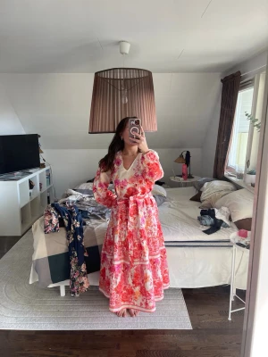 Blommig långklänning med volang - Långklänning med blommigt mönster i rosa, rött och vitt. Klänningen har v-ringning, långa ärmar med volangdetaljer och ett matchande knytbälte i midjan. Tillverkad i ett lätt och luftigt material, perfekt för varma dagar. storlek L, nypris 999kr