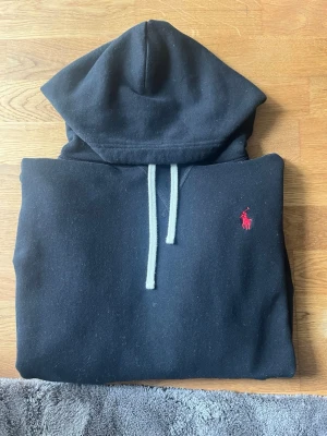 Svart hoodie från Polo Ralph Lauren - Säljer en svart hoodie från Polo Ralph Lauren med klassisk röd logga på bröstet. Hoodien har vit dragsko, stor huva och en magficka. Tillverkad i mjukt bomullsmaterial som är perfekt för chill dagar. Enkel och stilren design som funkar till det mesta.