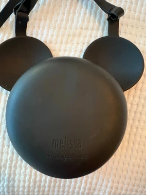 Svart handväska med musöron - Cool svart handväska i form av ett runt huvud med två stora öron, inspirerad av tecknade figurer. Väskan har dragkedja upptill och justerbar axelrem. Tillverkad i slätt konstläder som ger en stilren look.