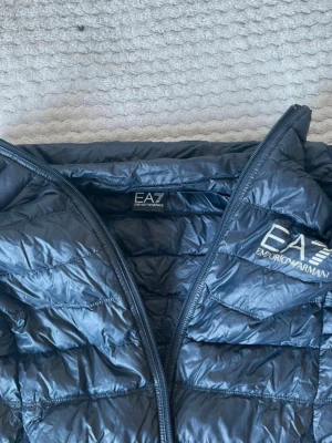 Svart dunväst från Emporio Armani EA7 - Snygg svart dunväst från Emporio Armani EA7 med quiltad design och logotyp på bröstet. Västen har en hög krage och dragkedja framtill. Perfekt för att lägga till ett extra lager och ge en sportig look. Betalning genom vanlig swish! 