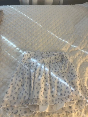 Vit blommig kortkjol från Gina Tricot - Söt vit kortkjol från Gina Tricot med blått blommönster. Kjolen har ett tunt, luftigt tyg och resår i midjan för skön passform. Perfekt för varma dagar och ger en fräsch och somrig look.