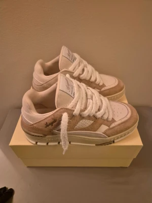Axel Arigato - Säljer ett par snygga sneakers från Axel Arigato i vitt och beige. Skorna har en låg siluett, breda vita snören och detaljer i mocka och läder. Märkeslogga på sidan och tungan. Perfekta för dig som gillar stilrena och trendiga sneakers.