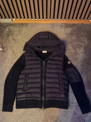 Moncler cardigan - Kom pm för bild på den på! Snygg svart pufferjacka från Moncler med stickade ärmar och huva. Jackan har quiltad front, två fickor med knappstängning och en extra ficka med dragkedja på ärmen. Ikonisk Moncler-logga på vänster ärm och dragkedja framtill.  