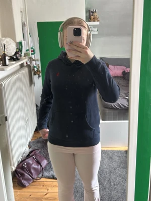 Mörkblå hoodie från Ralph Lauren - Säljer en mörkblå hoodie från Ralph Lauren med klassisk huva och känguruficka framtill. Diskret röd logga broderad på bröstet. Hoodien har en avslappnad passform och är tillverkad i mjukt material som känns skönt mot huden.