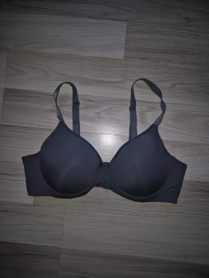 Mörkgrå vadderad bygel-bh - En enkel och stilren mörkgrå bh med vadderade kupor och bygel för extra stöd. Justerbara axelband och hakknäppning bak. Tillverkad i mjukt, slätt material som känns bekvämt mot huden. Perfekt för dig som vill ha en diskret och klassisk look.