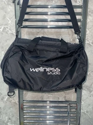 Svart träningsväska Wellness Studio - Snygg svart träningsväska från Wellness Studio med justerbar axelrem och bärhandtag. Väskan har dragkedja upptill och extra fack på sidan. Tillverkad i slitstarkt syntetmaterial, perfekt för gymmet eller sportaktiviteter.