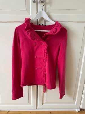 Rosa kofta med volangkrage - Snygg rosa kofta med dubbelknäppning och volangdetaljer längs kragen. Långärmad modell i mjukt tyg 