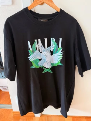 Svart Amiri t-shirt med duvor - Säljer en svart t-shirt från Amiri (REPLIKA!) med stort tryck av vita duvor och gröna blad på bröstet. T-shirten har rund hals, korta ärmar och en relaxed passform. Perfekt för dig som gillar streetwear och vill sticka ut med en unik design.