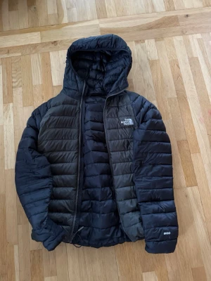 Svart dunjacka från The North Face - Snygg svart dunjacka från The North Face med huva och dragkedja framtill. Jackan har quiltad design, två sidofickor och logga på bröstet. Perfekt för kalla vinterdagar tack vare dunfyllningen och den lätta, smidiga passformen.