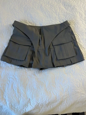 Grå skort med fickor och detaljer - Unik grå skort med stora fickor framtill och coola spetsiga detaljer vid midjan. Skorten har en modern, edgy look med flera lager och markerade sömmar. Perfekt för dig som vill sticka ut med din stil.