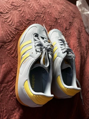 Adidas Samba sneakers ljusblå/gul - Säljer ett par Adidas Samba sneakers i ljusblå mocka och läder med gula detaljer och klassiska vita stripes. Yttersulan är i gummi med retro mönster. Skorna har låg profil och snörning framtill. Perfekta för dig som gillar sportig och stilren look.