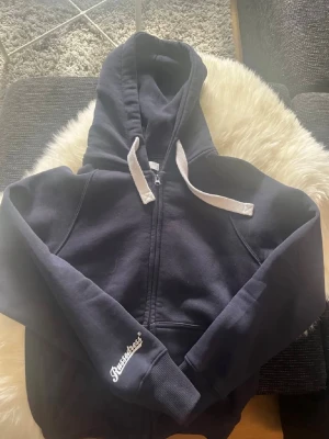 Mörkblå hoodie från Russemerch. - Mörkblå zip-hoodie från Russemerch med vit dragsko i huvan och broderad logga på ärmen. Klassisk passform med ribbade muddar och fickor framtill. Perfekt för en avslappnad och sportig stil. Säljer eftersom den inte kommer till användning. Perfekt nu till sommaren ifall det är lite kallt och man bara vill dra på något.