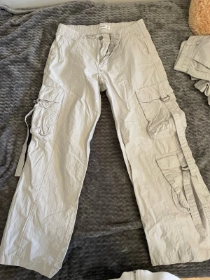 Pull&bear cargobyxor med fickor och band - Säljer ett par  cargobyxor med flera stora fickor på benen och dekorativa banddetaljer. Byxorna har avslappnad passform och är tillverkade i ett lätt bomullsliknande material. Perfekta för en streetwear-look.