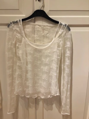 Vit spetsblus från SO Goods For Life - Säljer en vit långärmad blus från SO Goods For Life i storlek XS. Blusen är gjord i ett tunt, transparent spetsmaterial med blommigt mönster och har en rundad halsringning, med ett linne under. Använd ungefär 1 gång 