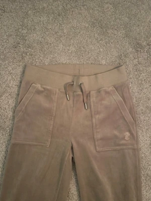 Beige Juicy Couture byxor  - Säljer ett par beige mjukisbyxor från Juicy Couture i mjuk velour. Byxorna har bred resår i midjan med dragsko, stora fickor fram och diskret JC-logga broderad på ena fickan. Perfekta för en chill och trendig look.