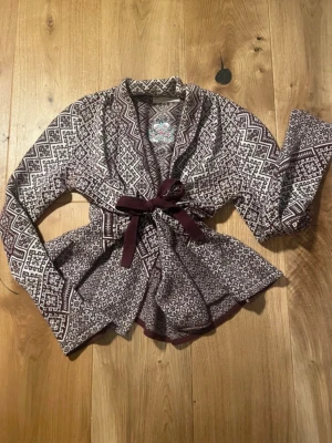 Odd Molly kofta med knytskärp - Äkta Odd Molly,  Snygg kofta i vinrött och vitt med grafiskt mönster och knytskärp i midjan. Koftan har en öppen design utan knappar. Perfekt för lager-på-lager och ger en cool touch till din outfit.