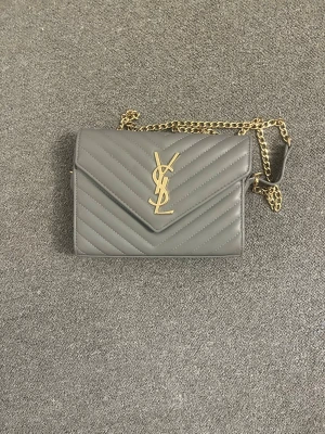 Grå YSL kuvertväska med kedja - Snygg grå kuvertväska från YSL i quiltat läder med guldfärgad kedjerem och ikonisk YSL-logga framtill. Väskan har ett klassiskt kuvertformat lock och elegant design som passar perfekt till alla tillfällen.