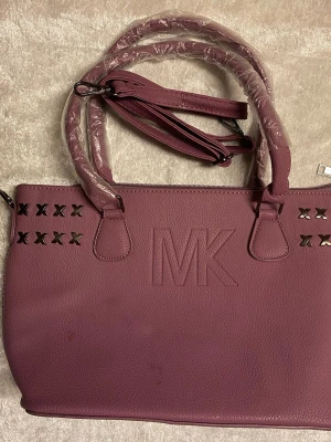Lila handväska  - Snygg lila handväska  i konstläder med präglad MK-logga framtill. Väskan har dekorativa metallkryss, dubbla handtag och avtagbar axelrem. Praktisk dragkedjeficka på baksidan och rymligt huvudfack. Perfekt accessoar för en trendig look.