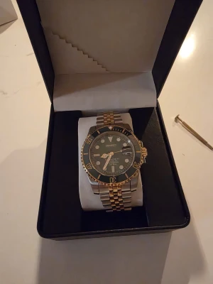 Seiko automatisk klocka silver/guld - Snygg automatisk klocka från Seiko med tvåfärgad länk i silver och guld. Svart urtavla med tydliga index och datumvisning. Boetten har gulddetaljer och glasbaksida som visar urverket. Perfekt accessoar för dig som gillar klassisk stil.