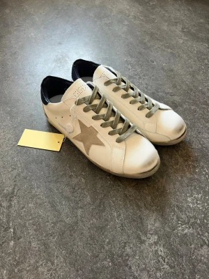 Vita sneakers med stjärndetalj - Tjena, säljer dessa snygga vita skor från Golden Goose med grå stjärna på sidan, grå skosnören och mörkblått hälparti. 