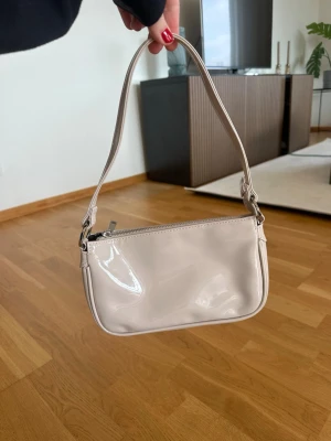 Beige axelremsväska i lack - Säljer en stilren beige axelremsväska i glansigt lackmaterial. Väskan har en enkel, rektangulär form med silverfärgade metalldetaljer och dragkedja upptill. Perfekt storlek för det viktigaste och ger en trendig touch till din outfit.