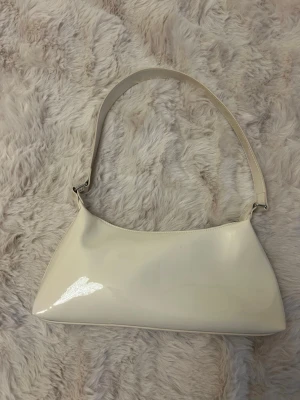 Vit axelremsväska i lack - Stilren vit handväska i glansigt lackmaterial med axelrem. Väskan har en avlång, mjukt triangulär form och stängs med dragkedja upptill. Perfekt för dig som gillar minimalistisk och trendig design.