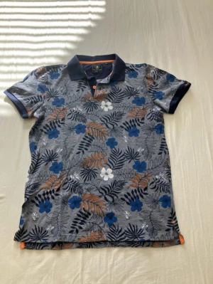 Blommig pikétröja från New Zealand - Snygg pikétröja med blommigt mönster i blått, vitt och orange på grå botten. Krage och ärmslut i mörkblått, med knappar framtill. Perfekt för en avslappnad och trendig stil.