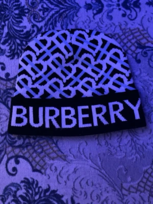 Svartvit Burberry mössa med logga - Säljer en svartvit mössa från Burberry med stort logotyp-tryck framtill och ett grafiskt mönster över hela mössan. Tillverkad i mjukt stickat material som håller dig varm och snygg. Perfekt accessoar för dig som gillar streetstyle och designerplagg.