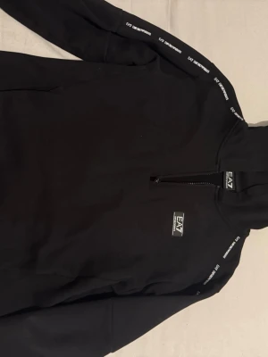 Svart hoodie från EA7 Emporio Armani - Snygg svart hoodie från EA7 Emporio Armani med dragkedja framtill och logga på bröstet. Tillverkad i mjukt bomullsmaterial som känns skönt mot huden. Perfekt för dig som gillar stilrena och sportiga plagg med exklusiv känsla. Klassisk huva och diskreta detaljer.